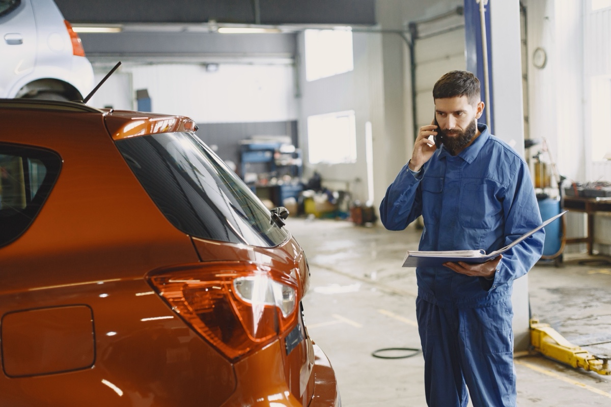 Profissional de uniforme azul falando ao telefone e segurando uma pasta ao lado de um carro dentro de uma oficina