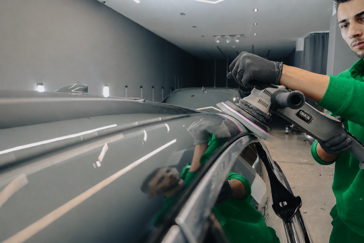 Profissional de camisa verde passando politriz no teto de um carro escuro dentro de um estúdio de estética automotiva