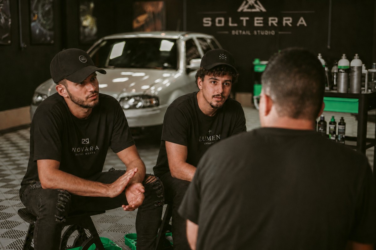 Três profissionais de estética automotiva conversando dentro de um estúdio de detalhamento, ao lado de um carro branco